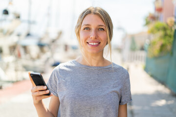 Young blonde woman using mobile phone at outdoors smiling a lot