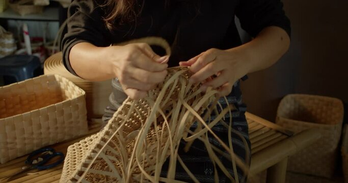 An Asian female artisan who is a rural villager weaving folk women's baskets and bags made from leftover paper band to sell as extra income in the community shop.