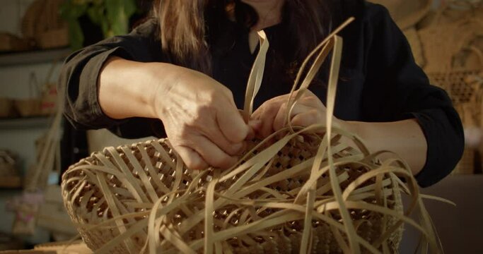 An Asian female artisan who is a rural villager weaving folk women's baskets and bags made from leftover paper band to sell as extra income in the community shop.