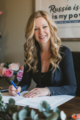 Fototapeta premium Gorgeous blonde Russian lady late thirties sitting office desk and signing papers, documents like contract, deal or agreement. Businesswoman in black business attire gives her signature.