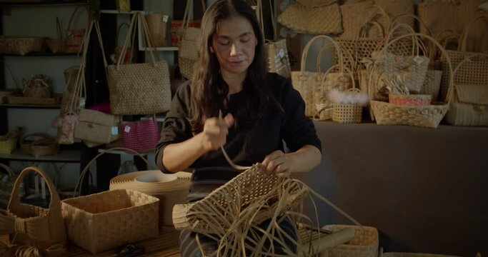 An Asian female artisan who is a rural villager weaving folk women's baskets and bags made from leftover paper band to sell as extra income in the community shop.