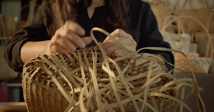 An Asian female artisan who is a rural villager weaving folk women's baskets and bags made from leftover paper band to sell as extra income in the community shop.