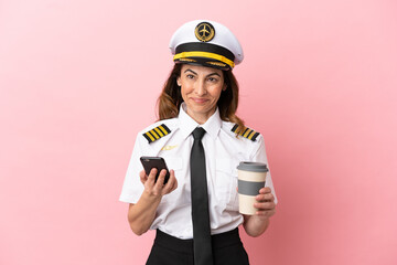 Airplane middle aged pilot woman isolated on pink background holding coffee to take away and a mobile