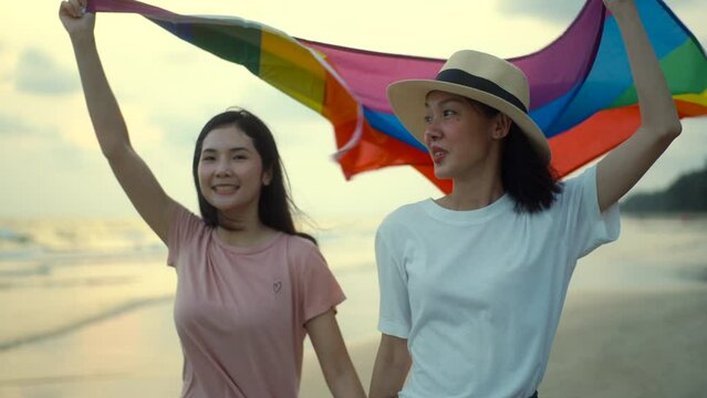 Bisexual lesbian girlfriend woman couple walking on the beach with their arms crossed with rainbow flag. LGBTQ female couple have a romantic moment on the beach. LGBT and homosexual concept.
