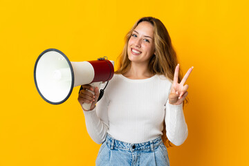 Young blonde woman isolated on yellow background holding a megaphone and smiling and showing...