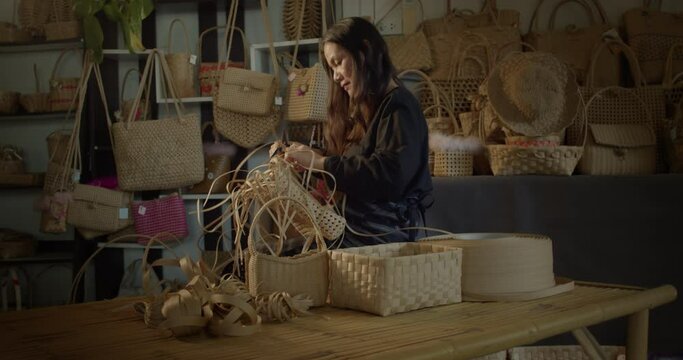 An Asian female artisan who is a rural villager weaving folk women's baskets and bags made from leftover paper band to sell as extra income in the community shop.