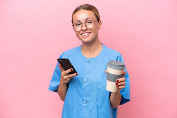 Young nurse doctor woman isolated on pink background holding coffee to take away and a mobile - 783628270