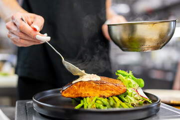 Professional chef preparing a dish of grilled salmon, asparagus, and fresh salad, drizzling sauce from a small bowl in a kitchen setting