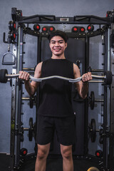 A young slim asian man in a black tank top does basic standing barbell curls at the gym. Using an...