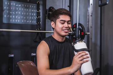 A young man enjoys a sip of cold water or sports drink from a insulated water bottle while sitting on a bench. Quenching thirst between workouts at the gym.