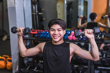 A smiling young man does a set of seated dumbbell shoulder presses. A slim teenager building muscle at the gym.
