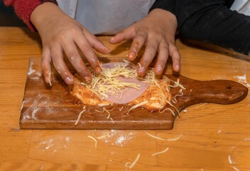 Young boy preparing ingredients for the pizza on the brown wooden board in the kitchen