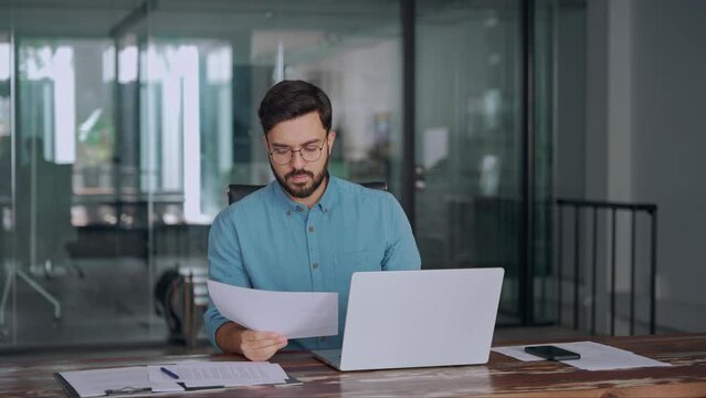 Busy Latin financial expert checking documents working at laptop in office. Serious business man accountant eading law paper company file invoice overview looking at computer at workplace.