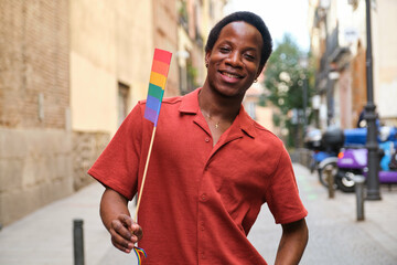 Gay black man in a red shirt is holding a rainbow flag and smiling at Pride Month.