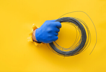 A righ man's hand in a blue knitted glove holds a coil of steel wire. Torn hole in yellow paper. The concept of a worker, a labor migrant, a master of his craft. Copy space. - Powered by Adobe
