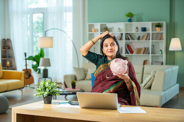 Fototapeta na wymiar Finance concept - Indian retired old lady holding piggy bank, money, laptop and 3d home model at home