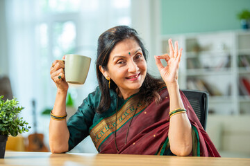 Happy beautiful relaxed mature older adult Indian asian woman drinking coffee or tea at home.