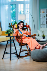 Old retired Indian woman sitting on rocking chair indoors with cup of coffee, tea or juice glass