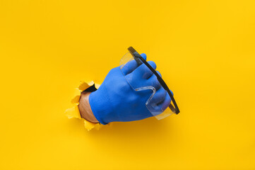 The right male hand in a blue fabric work glove holds safety glasses. Torn hole in yellow paper. Good job, eye protection and safety concept. Copy space.