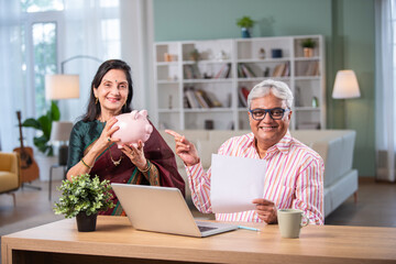 Indian asian retired couple accounting at home with laptop, papers, money and piggy bank