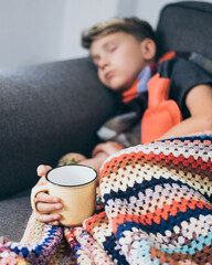 Sick boy sleeping with woolen blanket, hot water bottle and a mug. Sad teen with the flu rests alone at home in a cold winter day. Child with seasonal infections and fever Soft focus on the cup.