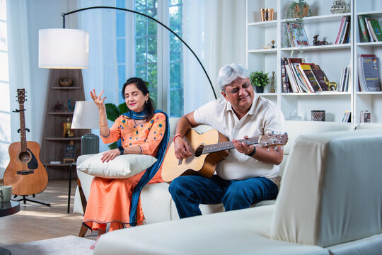 Indian asian senior husband playing acoustic guitar and wife singing, both sitting together on sofa.