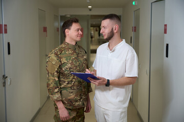 Physician making notes in hallway in presence of military patient - obrazy, fototapety, plakaty