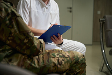 Medical professional writing on clipboard health complaints of serviceman - obrazy, fototapety, plakaty