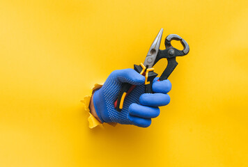 A right man's hand in a blue fabric knitted glove holds wire cutters and pliers. Torn hole in...