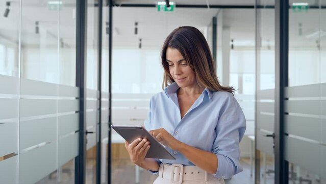 Busy middle aged businesswoman manager, older female executive leader holding pad digital tablet at work, looking at pad. Smiling mature professional business woman using tablet standing in office.