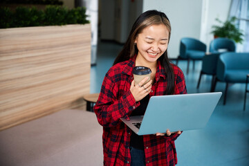 Laughing asian female IT employee watching laptop in hall of coworking office