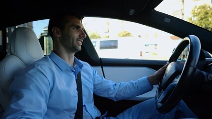 Profile of handsome businessman driving a modern electric car at urban road. Male successful entrepreneur riding on electrical vehicle at city. Young man commuting to work at sunny day. Slow motion