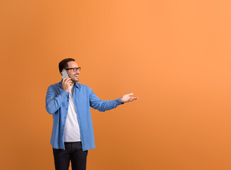 Happy young businessman talking on smart phone and showing empty palm on isolated orange background