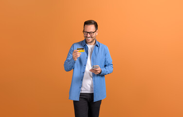 Smiling young businessman shopping online with smart phone and credit card over orange background