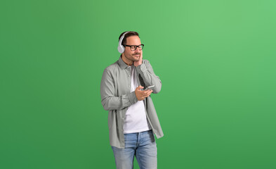 Smiling businessman with eyes closed listening music over headset and relaxing on green background