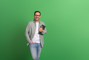 Portrait of happy entrepreneur with hand in pocket holding laptop over isolated green background
