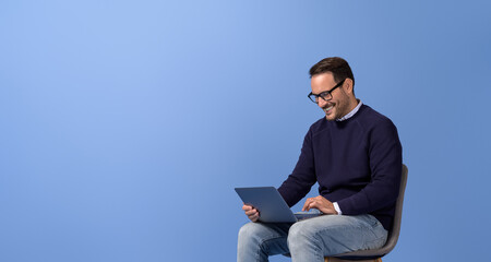 Smiling businessman doing online research over wireless computer on chair against blue background