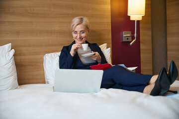 Smiling businesswoman drinking tea or coffee from cup and watching laptop on bed