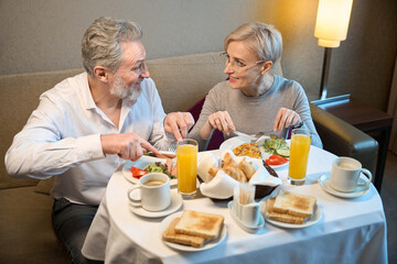 Smiling adult caucasian couple having breakfast and looking at each other - obrazy, fototapety, plakaty