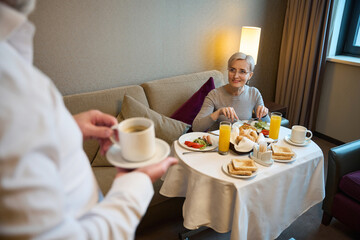Woman having breakfast and looking at cropped man with cup of tea or coffee
