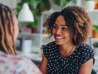 Two women are sitting at a table, one of them smiling. The woman on the left is wearing a floral shirt and the woman on the right is wearing a black polka dot shirt. They are talking to each other - obrazy, fototapety, plakaty