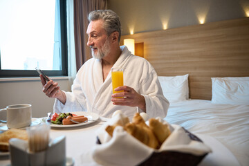 Caucasian man watching smartphone and drinking juice during having breakfast