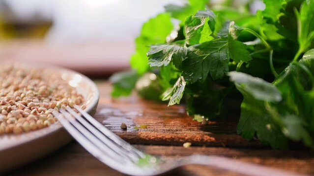 green parsley and cilantro in a bunch falls on the kitchen table. Next to the table there is a fork and a plate with buckwheat. High quality 4k footage