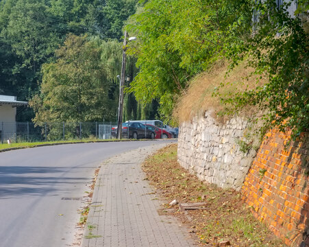 Fototapeta Jesień na ulicach miasta - mur z cegły i kamienia. Suche jesienne liście na chodniku na ulicy miejskiej wśród wielu drzew.