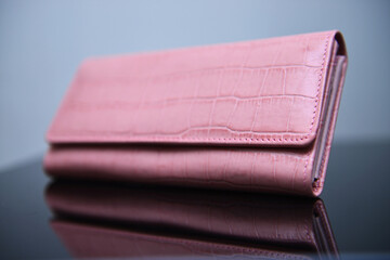 A pink color ladies' purse made with premium quality leather of crocodile skin texture, placed on the glass table and with its reflection
