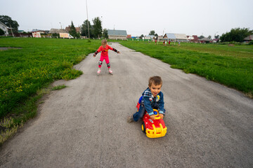 a boy in a denim jacket sits on a toy car, a girl roller-skates, happy children, sister and brother, family and sports, toy car, spring, walk after the rain	