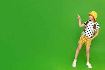 A child in summer clothes points to your ad. A young girl in shorts and a hat is enjoying her summer vacation. Children's holidays. Green isolated background. Copy space. Place for the text, banner
