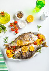 Sea bream fish dorado with tomato cherries and lemon slices on white plate. Top view.