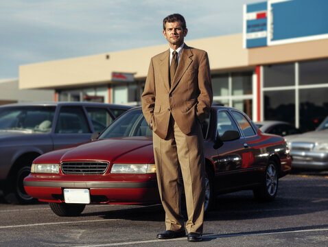 A sleazy car salesman in a polyester suit stands in his lot, ready to pressure customers.
