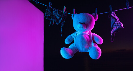A toy teddy bear in a bdsm mask and leather straps hangs on a clothesline in neon light on a dark...